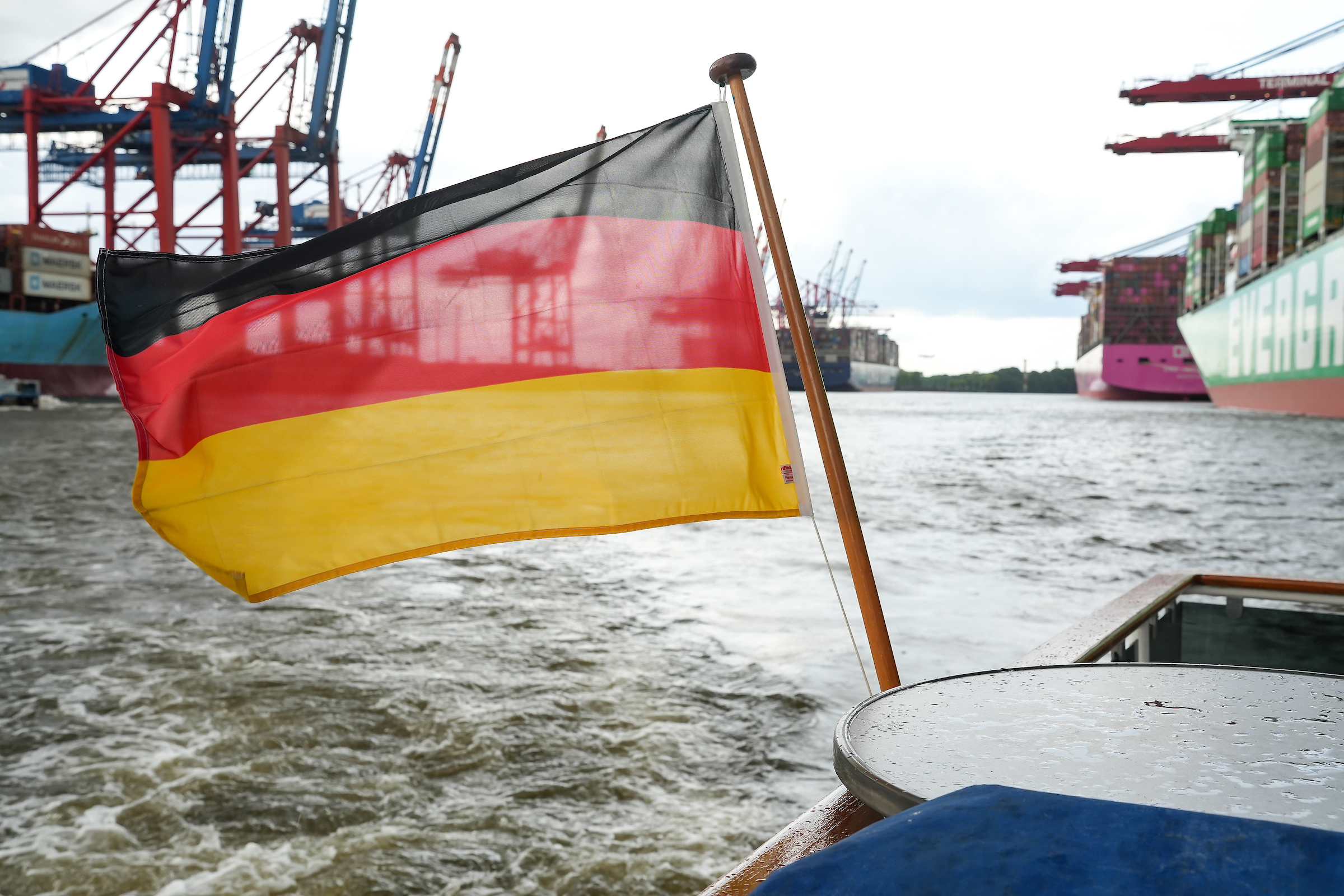 German flag on a boat in the Port of Hamburg, with shipping cranes and container ships in the background.